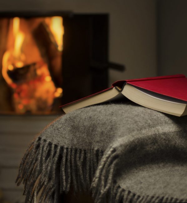 Peceful image of open book resting on a arm rest of a couch. Warm fireplace on background.